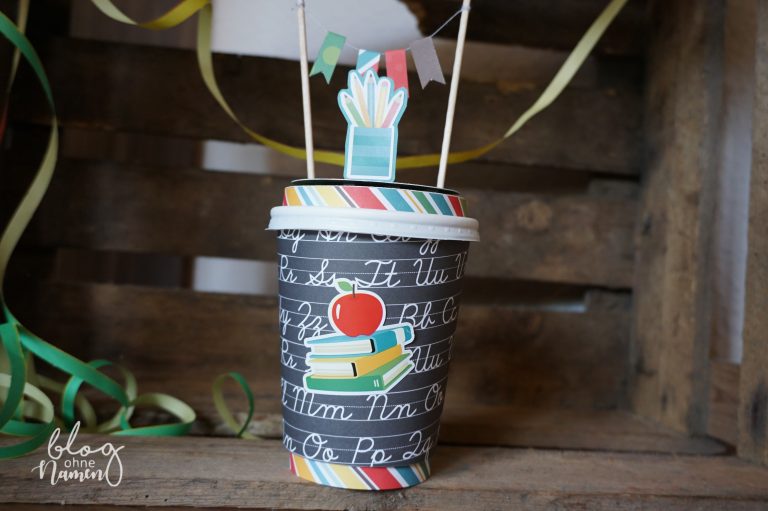 Einschulungsgeschenk mal anders: Ein gestalteter Coffee-to-go-Becher mit passender Banderole und Schuldesign-Papieren als originelle Verpackungsidee.